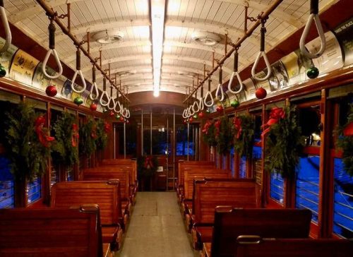 Interior of a vintage train car decorated with holiday wreaths and lights, featuring wooden benches and a warm ambiance.