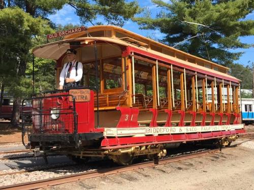 A vintage red trolley car with a conductor stands on the tracks surrounded by trees.