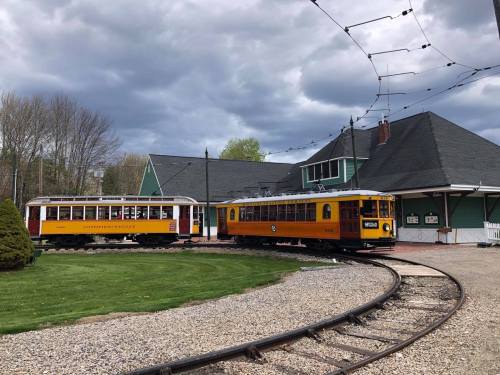 Two vintage trolleys in yellow and orange parked near a green building, with a cloudy sky overhead.
