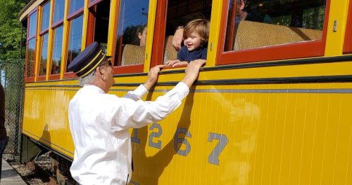 A conductor interacts with a smiling child leaning out of a vintage train window. Bright yellow train car in the background.
