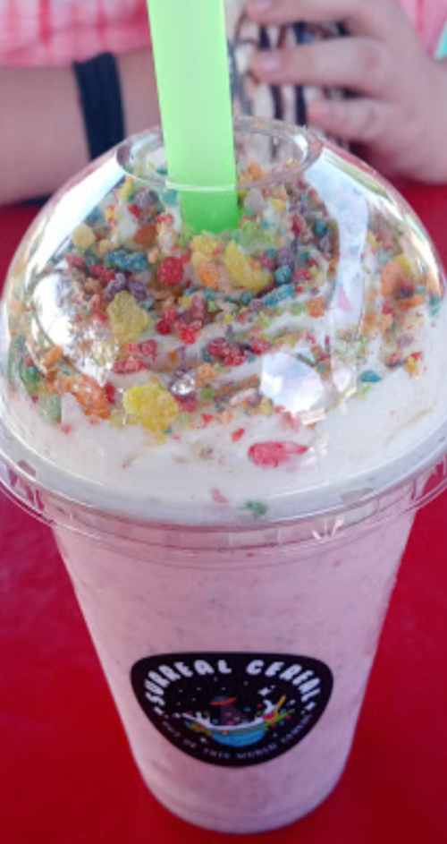 A colorful milkshake topped with cereal and a green straw, served in a clear cup with a logo.