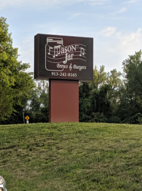 Sign for "Jason Jar Brews & Burgers" with a musical theme, located on a grassy area with trees in the background.