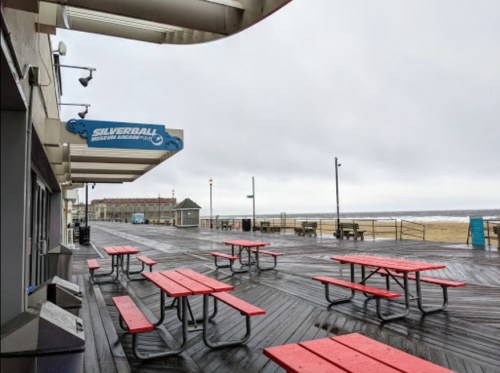 Empty boardwalk with red picnic tables, a closed arcade, and a cloudy sky over the beach.
