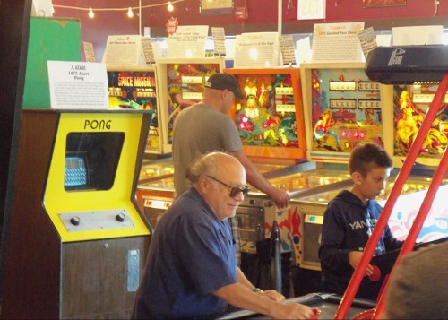 A retro arcade with pinball machines, a man playing a game, and a boy observing, all in a vibrant, nostalgic setting.