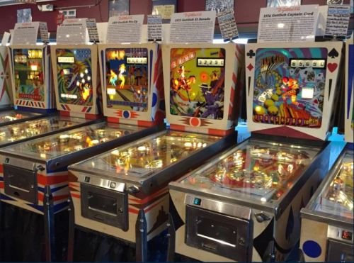 A row of vintage pinball machines with colorful designs and illuminated playfields in a retro arcade setting.
