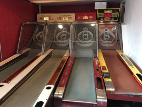 Four vintage skee ball machines lined up against a red wall, featuring scoring holes and wooden lanes.