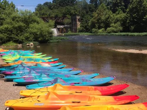 Colorful kayaks lined up along a riverbank with a dam and lush greenery in the background.