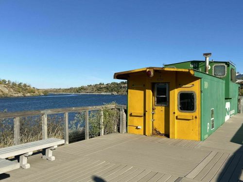 A yellow and green train car on a wooden deck by a calm river under a clear blue sky.