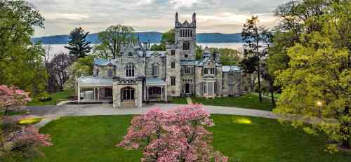 A grand, historic mansion surrounded by lush greenery and blooming trees, overlooking a serene body of water.