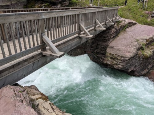 A wooden bridge spans a rushing river, surrounded by rocky terrain and greenery.