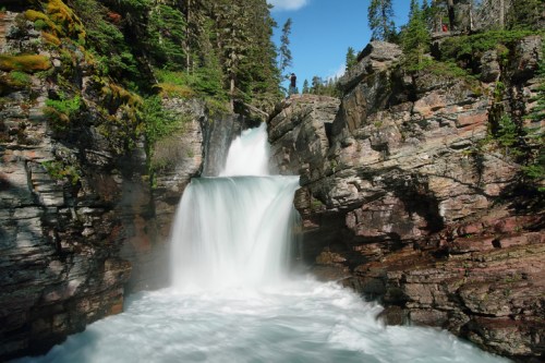 A serene waterfall cascades over rocky cliffs, surrounded by lush greenery and tall trees under a clear blue sky.