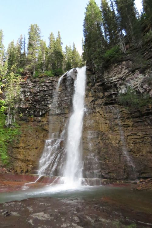 A tall waterfall cascades down rocky cliffs, surrounded by lush green trees and a clear blue sky.