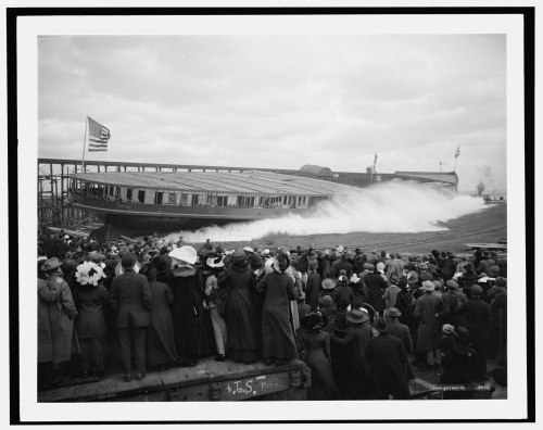A large crowd watches as a ship is launched into the water, with waves splashing against its hull.
