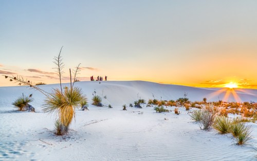 A sunset over white sand dunes, with silhouettes of people on the horizon and sparse vegetation in the foreground.