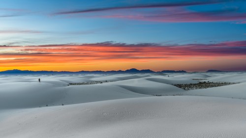 A serene landscape of white sand dunes under a colorful sunset sky with distant mountains.