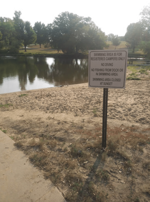 Sign by a sandy area near a calm lake, stating swimming rules and restrictions for registered campers only.