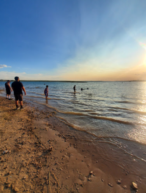 People wade in shallow water at a beach during sunset, with a clear sky and gentle waves.