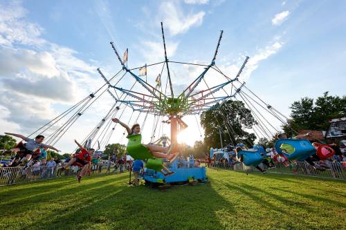 Children enjoy a swing ride at a fair, with colorful flags and a sunset in the background.