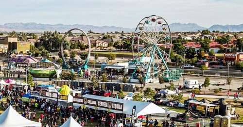Aerial view of a lively fair with rides, food stalls, and crowds, set against a backdrop of mountains and buildings.