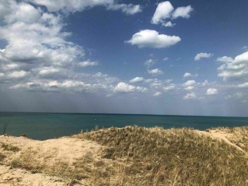 A sandy dune with grass overlooks a calm blue sea under a partly cloudy sky.