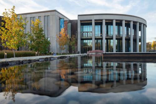 Modern building with large glass windows, surrounded by trees, reflecting in a calm water surface.