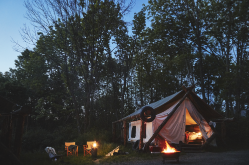 A cozy tent in a forest at dusk, with a fire pit and chairs nearby, surrounded by trees.
