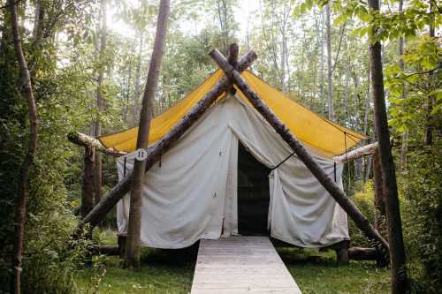 A canvas tent with a yellow roof, set in a forest, surrounded by trees and a wooden walkway leading to the entrance.