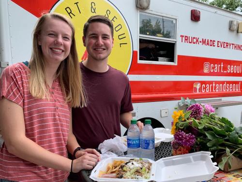 A smiling couple stands by a food truck, holding a takeout container and drinks, with flowers on the table.