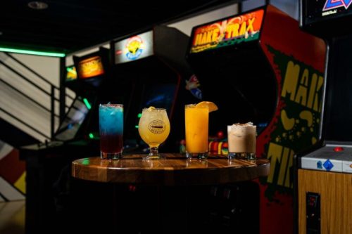 Four colorful cocktails on a wooden table in front of vintage arcade machines.