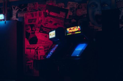A dimly lit arcade with a vintage Frogger machine surrounded by colorful posters on the wall.