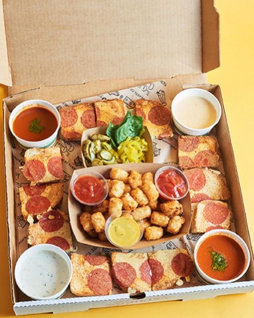 A pizza box filled with pepperoni pizza slices, tater tots, pickles, and various dipping sauces.