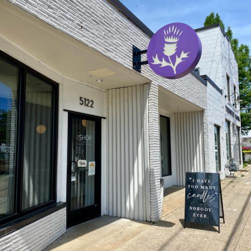 Exterior of a shop with a purple thistle sign, featuring a humorous quote about candles on a sidewalk sign.