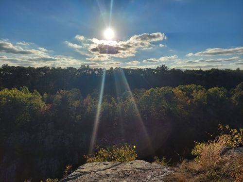 A sunlit landscape with a bright sun, clouds, and colorful trees in the foreground and a deep canyon in the background.