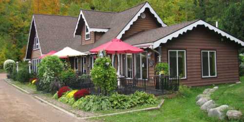 A charming brown house with a sloped roof, surrounded by colorful flowers and greenery, featuring outdoor seating areas.