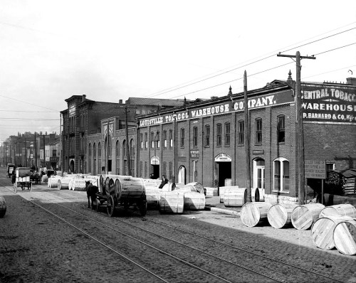 Historic black-and-white photo of a tobacco warehouse with barrels and horse-drawn carts on a cobblestone street.