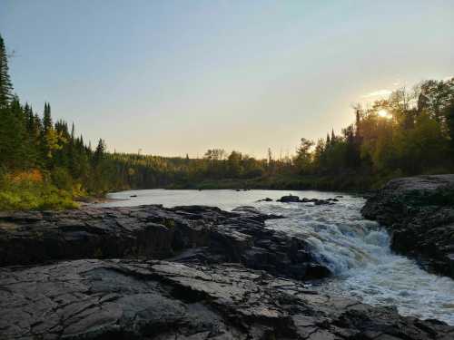 A serene river flows over rocky terrain, surrounded by lush trees and bathed in warm sunlight during sunset.