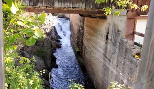 A wooden bridge spans a rocky area with a waterfall cascading into a narrow, flowing river below. Green foliage surrounds the scene.