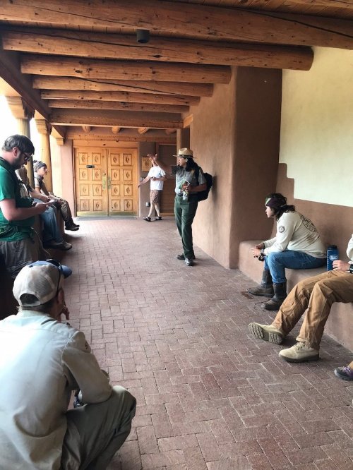 A group of people seated in a corridor, listening to a speaker standing near a wooden door.