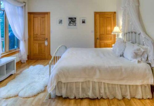 Cozy bedroom with a white bed, wooden doors, and a soft rug on a hardwood floor, featuring natural light from a window.