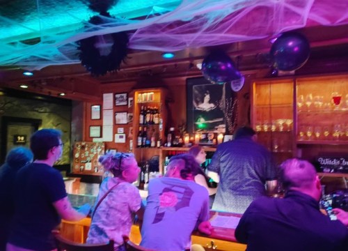 A dimly lit bar scene with patrons at the counter, decorated with spider webs and balloons for a festive atmosphere.