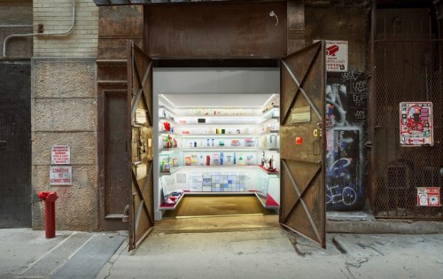 A modern store entrance with open double doors, showcasing colorful products on shelves inside. Urban setting with graffiti.