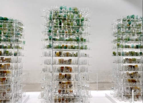 Three transparent structures resembling trees, filled with various green and brown glass containers, displayed on a white surface.