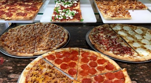 A variety of pizzas displayed on trays, including pepperoni, BBQ chicken, and veggie options.