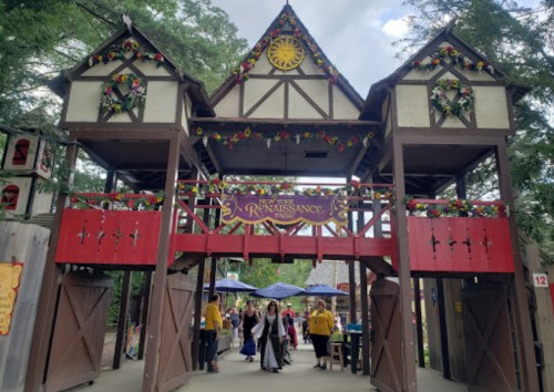 Entrance to a Renaissance fair, featuring a wooden archway adorned with flowers and a sign, with visitors in costumes.