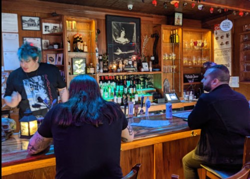 A bartender serves drinks at a wooden bar while two patrons sit, engaged in conversation. The bar is well-stocked with bottles.