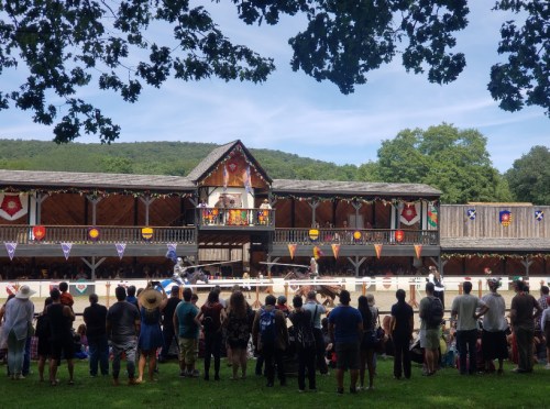 A crowd watches a performance at a medieval-themed festival, with colorful decorations and a wooden stage in the background.