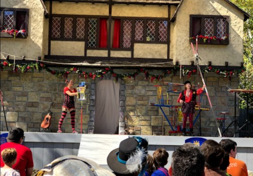 Two performers in colorful costumes entertain an audience on a stage decorated with flowers and a medieval-style backdrop.
