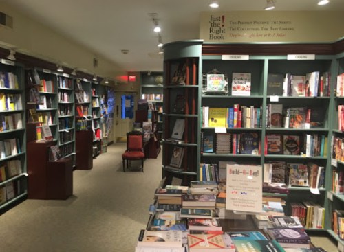A cozy bookstore interior with shelves of books, a table displaying titles, and a red chair in the background.
