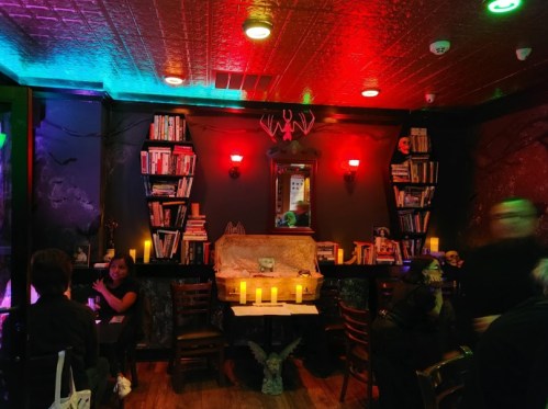 A dimly lit bar with colorful lights, bookshelves, candles, and a decorative coffin, creating a spooky atmosphere.