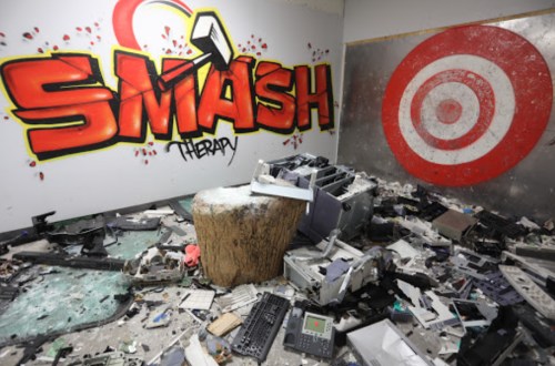 A messy room with broken electronics, a wooden stump, and graffiti reading "SMASH THERAPY" on the wall.